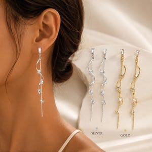 May include: Elegant silver and gold dangle earrings with a flowing, curved design. Each earring features sparkling clear crystals and a rectangular top. The image shows the earrings in silver and gold, with the word "SILVER" and "GOLD" below.