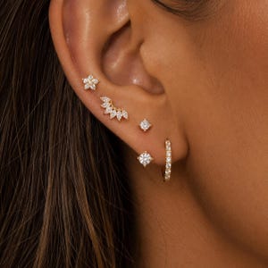 Puede incluir: Una colección de pendientes dorados con piedras preciosas transparentes. Los pendientes incluyen un pendiente de flor, un trepador de oreja curvado, dos pendientes cuadrados y un pequeño pendiente de aro.