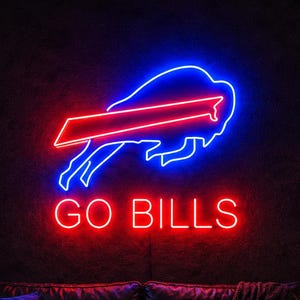 Puede incluir: Un letrero de neón montado en la pared con el contorno del logotipo de los Buffalo Bills en azul y rojo sobre el texto de neón rojo GO BILLS. El letrero emite un brillo intenso contra una pared oscura, sobre un sofá de cuero negro.