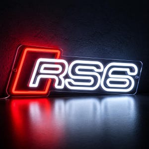 May include: A rectangular neon sign featuring a bright red outline of the Audi logo on the left side, followed by the white illuminated text RS6. The sign is mounted on a clear acrylic backing and emits a vibrant glow against a dark background.