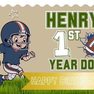 May include: A rectangular banner featuring a cartoon football player in a blue helmet and jersey, the text "HENRY'S 1ST YEAR DOWN", a football graphic, blue star outlines, and a gold ribbon banner with the text "HAPPY BIRTHDAY HENRY!" on green grass.