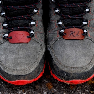 Può includere: Un paio di sneakers in camoscio grigio con lacci neri e rossi. Ogni scarpa presenta una targhetta in pelle marrone sui lacci; una è incisa con un simbolo a forma di cuore, l'altra con le lettere KL. Le sneakers hanno una suola in gomma rossa.