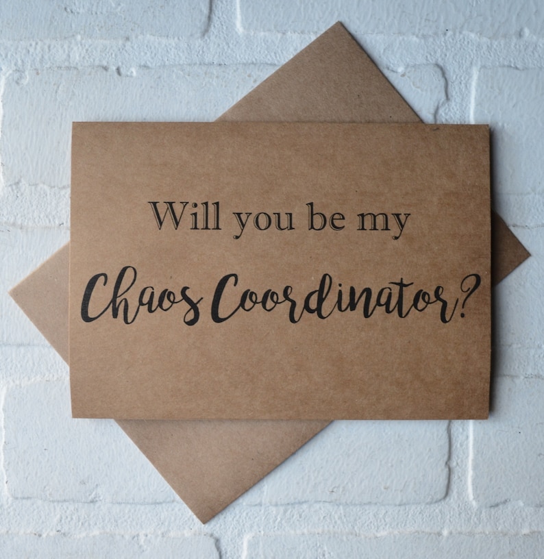 May include: A brown card with the text "Will you be my Chaos Coordinator?" in black lettering, resting on a brown envelope. The card and envelope are set against a white brick wall.
