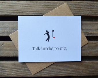 May include: A white card with a black silhouette of a golfer and a red heart. The text on the card reads "Talk birdie to me."