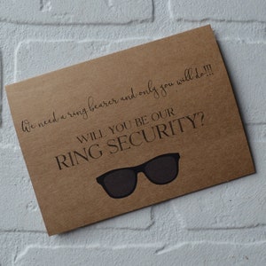 May include: A brown card with the text "We need a ring bearer and only you will do!!! Will you be our ring security?" and a pair of sunglasses.