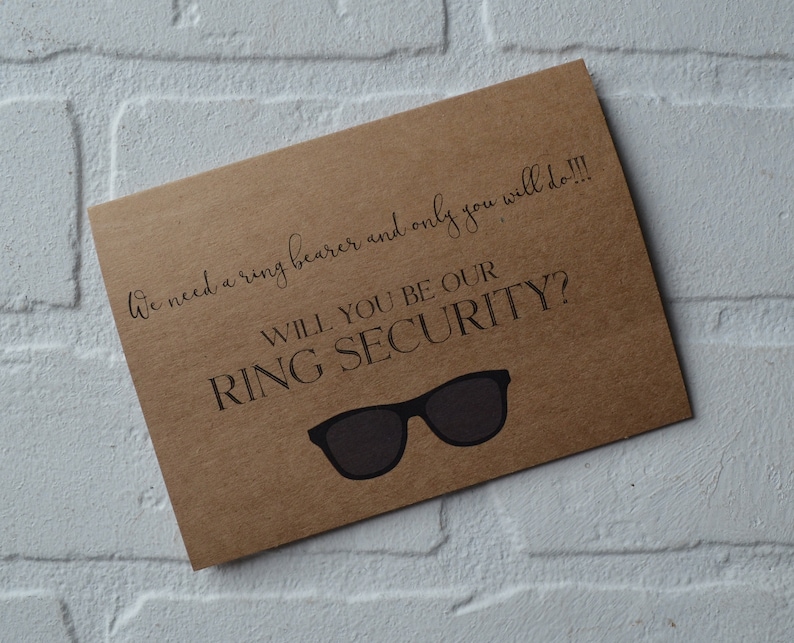 May include: A brown card with the text "We need a ring bearer and only you will do!!! Will you be our ring security?" and a pair of sunglasses.