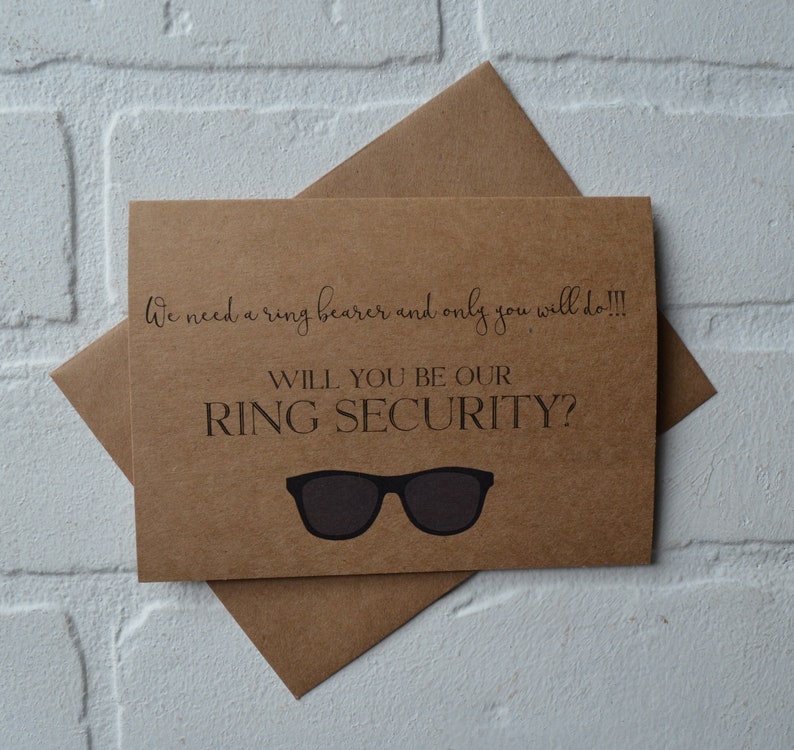 May include: A brown card with the text "We need a ring bearer and only you will do!!! Will you be our ring security?" with a pair of sunglasses drawn on the card.