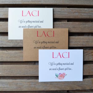 May include: Three white cards with the name "LACI" printed in pink letters. The cards have the text "We're getting married and we need a flower girl too..." printed in black. The bottom card has a watercolor floral design.