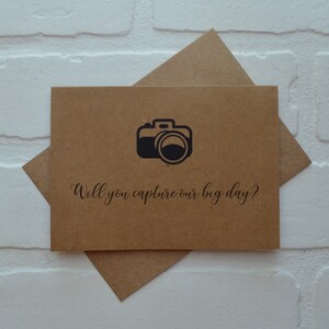 May include: A brown card with a black camera icon and the text "Will you capture our big day?" printed on it.