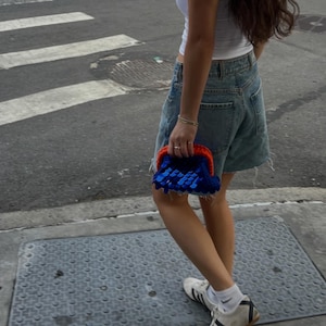 Puede incluir: Un bolso de mano pequeño y rectangular con un marco de ganchillo de color naranja vibrante y un cuerpo cubierto de lentejuelas brillantes de color azul intenso. El bolso se sostiene de lado, mostrando su diseño compacto y el contraste.
