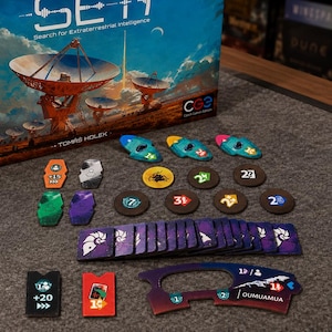 May include: A board game box titled SETI: Search for Extraterrestrial Intelligence featuring satellite dishes, accompanied by various game components including colorful tokens, circular markers, rectangular cards, and a shaped player board piece.