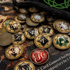 May include: A collection of round metal game tokens with a brass-toned finish, featuring various printed symbols, numbers, and icons including a skull, a green hood, and a red emblem. The tokens are scattered on a surface near a black fabric bag.