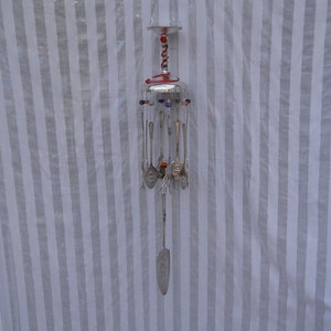 May include: A handcrafted wind chime made from repurposed silverware. The chime features a silver metal frame with hanging spoons, forks, and a serving utensil. Colorful beads and a glass top add decorative accents. The wind chime hangs from a black cord.