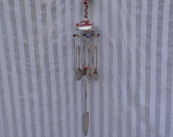 May include: A handcrafted wind chime made from repurposed silverware. The chime features a silver metal frame with hanging spoons, forks, and a serving utensil. Colorful beads and a glass top add decorative accents. The wind chime hangs from a black cord.
