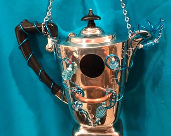May include: A silver-toned metal teapot repurposed as a birdhouse, featuring a circular opening on the front, a black handle, and a copper ring accent. The teapot is decorated with bright blue wire wrapping and assorted blue beads, suspended by a silver chain.