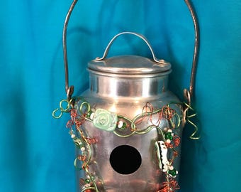 May include: A silver-colored metal milk can birdhouse with a circular entrance hole. The exterior is decorated with coiled green and copper-colored wire, small green and white beads, and a light green carved floral accent piece.