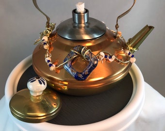 May include: A copper tea kettle with a brass handle and lid, decorated with white and blue beads and a blue and gold patterned ceramic charm. The kettle is sitting on a round, gray and white base.