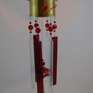 May include: A wind chime made from recycled materials. The chime features a brass bell, red glass beads, and clear glass panels. A silver spoon hangs from the bottom of the chime.