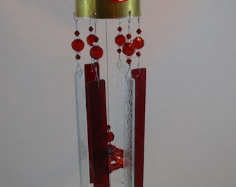 May include: A wind chime made from recycled materials. The chime features a brass bell, red glass beads, and clear glass panels. A silver spoon hangs from the bottom of the chime.