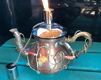 May include: A copper teapot with a silver lid and a flame coming out of the spout. The teapot is decorated with leaves and beads.