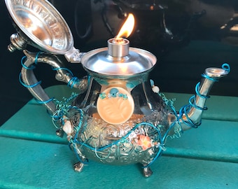 May include: A silver teapot with a teal blue wire design and a lit candle inside. The teapot has a floral design and is sitting on a teal blue surface.