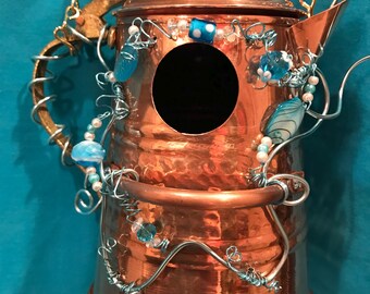 May include: A copper-colored metal teapot repurposed as a birdhouse, featuring a circular entry hole, a gold-toned chain for hanging, and decorative light blue wire accents adorned with various blue and white glass beads wrapped around the exterior.