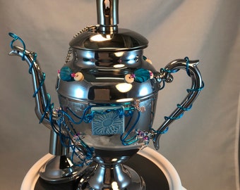 May include: A silver teapot with a blue and white floral design and a lit wick on top. The teapot is decorated with blue wire and beads.