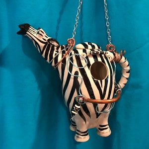 Könnte beinhalten: Eine Keramik-Teekanne in Form eines Zebras mit schwarzen und weißen Streifen. Die Teekanne hat ein Loch für Vögel, um hineinzufliegen, und eine Metallkette zum Aufhängen.