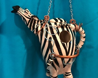 May include: A ceramic teapot shaped like a zebra with black and white stripes. The teapot has a hole for birds to enter and a metal chain for hanging.