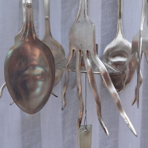 May include: A collection of silver-colored spoons and forks, some with unique, wavy tines, hanging against a white striped background. The utensils are arranged in a decorative display, showcasing their reflective surfaces.