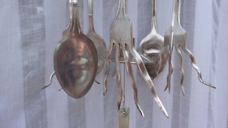 May include: A collection of silver-colored spoons and forks, some with unique, wavy tines, hanging against a white striped background. The utensils are arranged in a decorative display, showcasing their reflective surfaces.