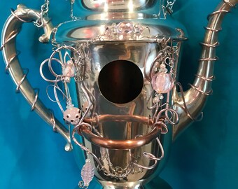May include: A silver-toned metal teapot repurposed as a birdhouse, featuring a circular entry hole, a copper-colored wire handle, and decorative pink glass beads wrapped in silver wire around the spout, handle, and body, suspended by a metal chain.