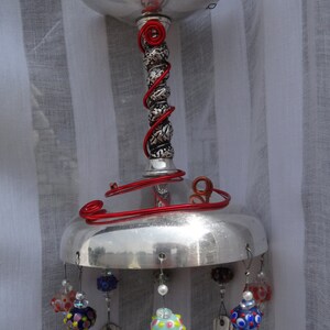 May include: A handcrafted wind chime featuring a silver-colored metal top and base, connected by a decorative stem wrapped with red wire. Colorful glass beads and repurposed silverware hang from the base, creating a unique and artistic outdoor decoration.