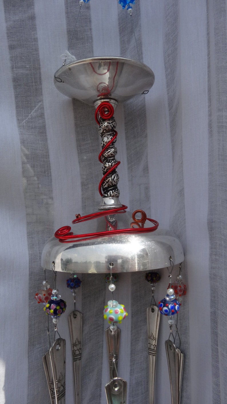 May include: A handcrafted wind chime featuring a silver-colored metal top and base, connected by a decorative stem wrapped with red wire. Colorful glass beads and repurposed silverware hang from the base, creating a unique and artistic outdoor decoration.