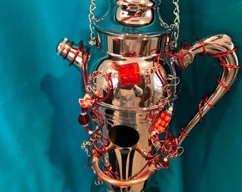 May include: A polished silver-tone metal cocktail shaker suspended by a silver chain. The shaker is wrapped in decorative red wire and adorned with various red and orange beads, including a prominent square red bead and a small floral accent.