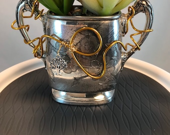 May include: A silver vintage creamer with ornate floral design, repurposed as a planter, holding a variety of succulents. The creamer has two handles with gold wire wrapped around them. A small silver charm with a tree design hangs from the wire.