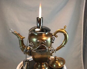 May include: A gold teapot with a silver lid and spout, decorated with a silver chain and black and white beads. The teapot has a small silver bell hanging from it and the word "CREATE" is written on the side. The teapot is lit with a flame coming from the spout.