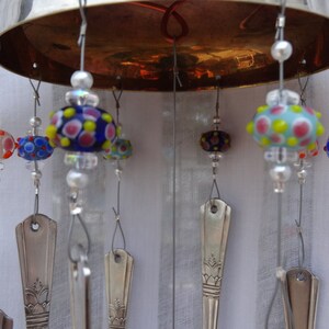 May include: A handmade wind chime featuring a silver-colored metal bowl top. Colorful glass beads with polka dot patterns in red, blue, and yellow are suspended from the bowl. Silverware spoons hang below the beads.