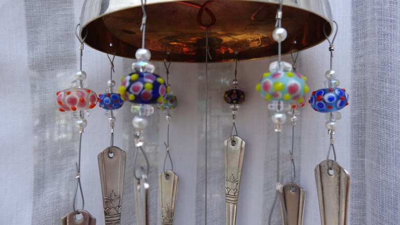 May include: A handmade wind chime featuring a silver-colored metal bowl top. Colorful glass beads with polka dot patterns in red, blue, and yellow are suspended from the bowl. Silverware spoons hang below the beads.