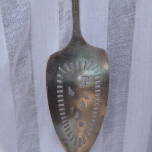 May include: Silver-toned serving spoon with a slotted design. The spoon has a wide, oval head with cut-out patterns and a long handle. The word "COMMUNITY" is engraved on the handle. The spoon is likely vintage or antique.