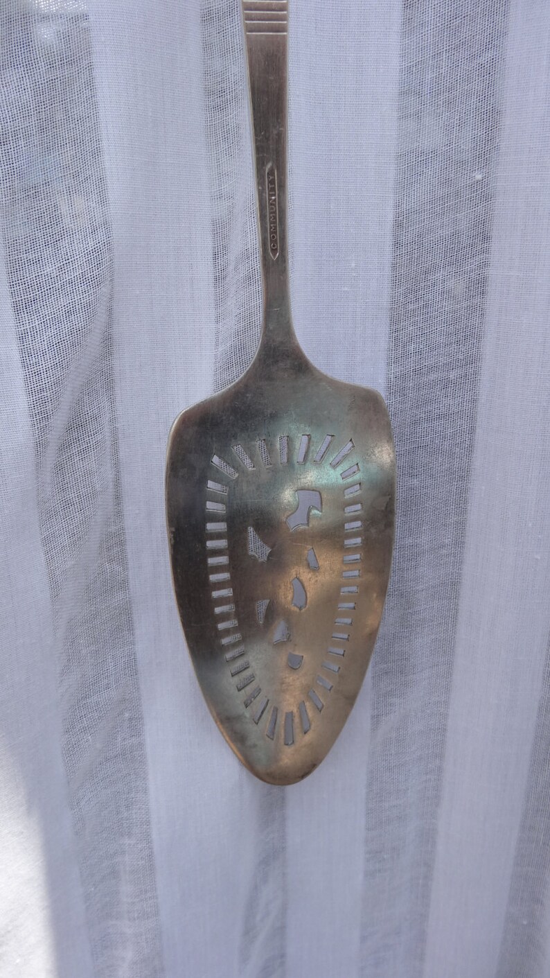 May include: Silver-toned serving spoon with a slotted design. The spoon has a wide, oval head with cut-out patterns and a long handle. The word "COMMUNITY" is engraved on the handle. The spoon is likely vintage or antique.
