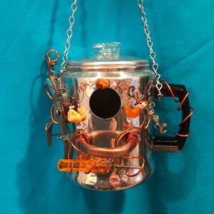 Könnte beinhalten: Ein silberner Kaffeebereiter aus Metall, der als Vogelhaus umfunktioniert wurde, mit einer runden Eingangsöffnung, einem klaren Glasdeckelknopf und einem schwarzen Griff. Verziert mit Kupferdraht, kleinen orangefarbenen Steinen und Werkzeugen.