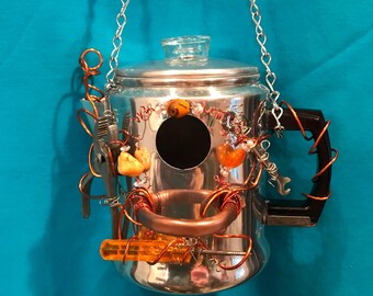 May include: A silver metal coffee percolator repurposed as a birdhouse, featuring a circular entrance hole, a clear glass lid knob, and a black handle. It is decorated with copper wire, small orange stones, a metal wrench, and an orange screwdriver.