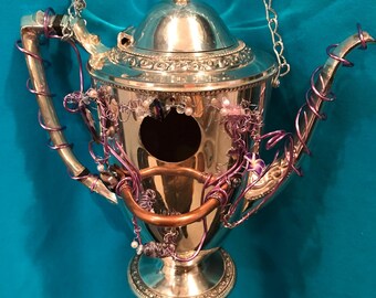 May include: A silver-toned metal teapot repurposed as a birdhouse, featuring a circular opening on the front. The teapot is adorned with purple wire coils, clear beads, and a copper-colored ring, and is suspended by a silver-toned metal chain.