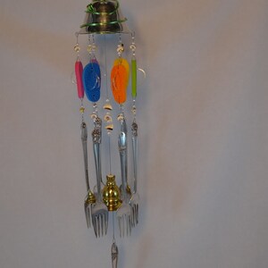 May include: A colorful wind chime made from recycled silverware, seashells, and flip-flops. The chime features a silver bell at the top and hangs from a silver chain.