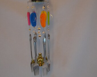 May include: A colorful wind chime made from recycled silverware, seashells, and flip-flops. The chime features a silver bell at the top and hangs from a silver chain.