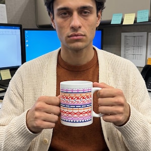 May include: A white ceramic mug featuring a horizontal band of colorful geometric patterns in shades of red, orange, pink, blue, and purple. The mug has a simple cylindrical shape with a curved handle on the right side, held by two hands.