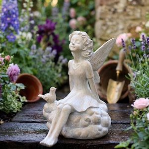 Könnte beinhalten: Eine Gartenstatue einer Fee, die auf einer Wolke sitzt, mit einem Vogel daneben. Die Fee hat zarte Flügel und trägt ein fließendes Kleid. Die Statue ist von bunten Blumen und Grün umgeben.