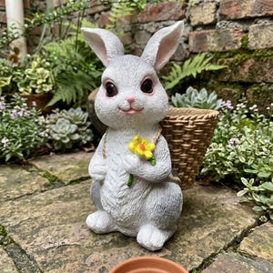 Puede incluir: Una figura de conejo gris claro con grandes ojos oscuros y nariz rosa, sosteniendo una flor amarilla en sus patas delanteras. El conejo lleva una cesta tejida de color canela en la espalda, asegurada con una cuerda, con pelaje texturizado.