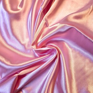 May include: A smooth, lustrous fabric with a reflective surface in shades of soft pink and light peach. The material is draped in deep, flowing folds that create a play of light and shadow, highlighting the glossy texture of the textile.